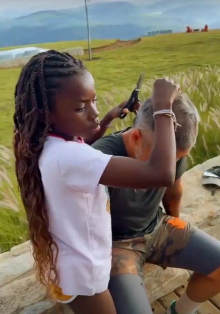 Titi de camisa branca, em pé, cortando o cabelo de Bruno Gagliasso, que está sentado