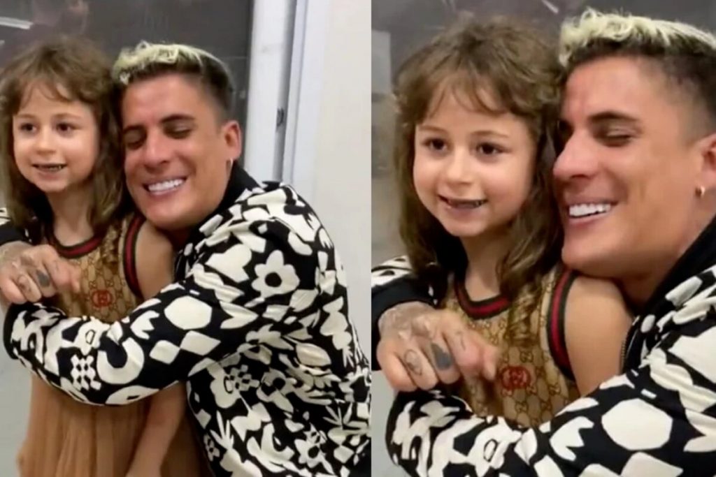 Tiago Ramos de casaco preto e branco, sorrindo, agachado no chão, abraçando Valentina Bezerra