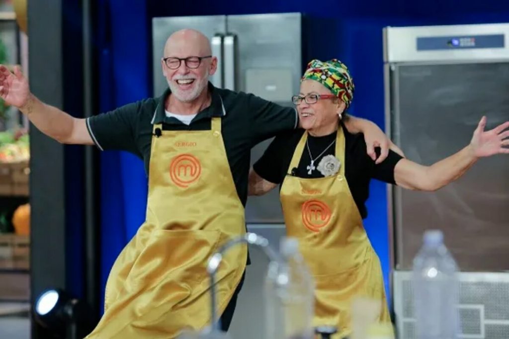 Sergio e Nadja, de avental dourado, abraçados e de braços abertos