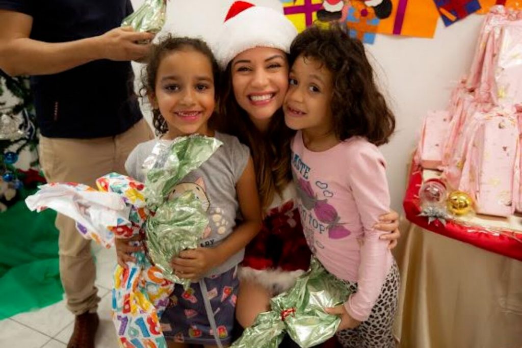 Maria Melilo vestida de Mamãe Noel, sentada com uma abraçada a duas meninas