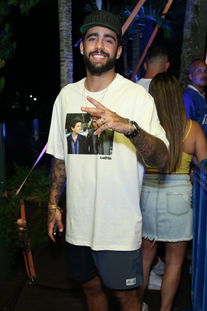 Pedro Scooby de camisa branca estampada, fazendo sinal de positivo e sorrindo