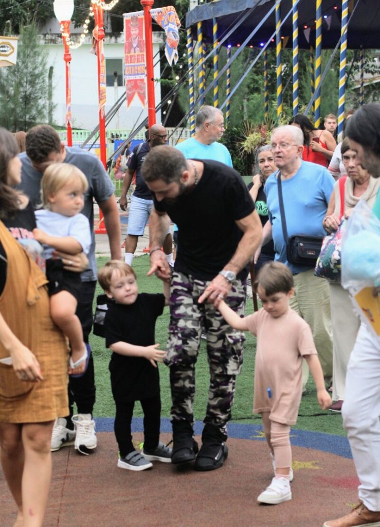 Thales Bretas de camisa preta, calça camuflada, de mãos dadas com os filhos, chegando ao circo