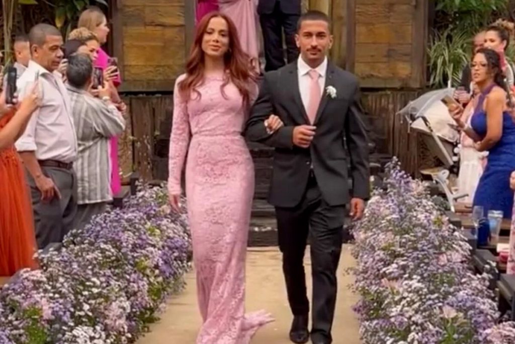 Anitta de vestido longo rendado, de manga longa, de braços dados com o irmão Renan