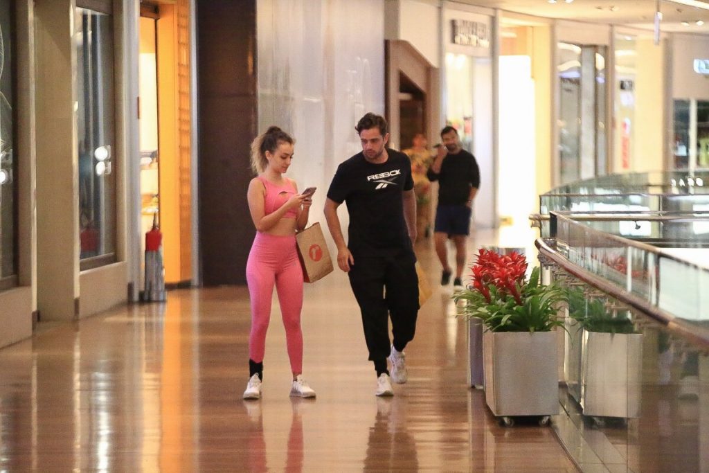 Rafael Cardoso de camisa preta e a namorada, Vivian Linhares, de top e legging rosa na escada rolante caminhando no corredor do shopping