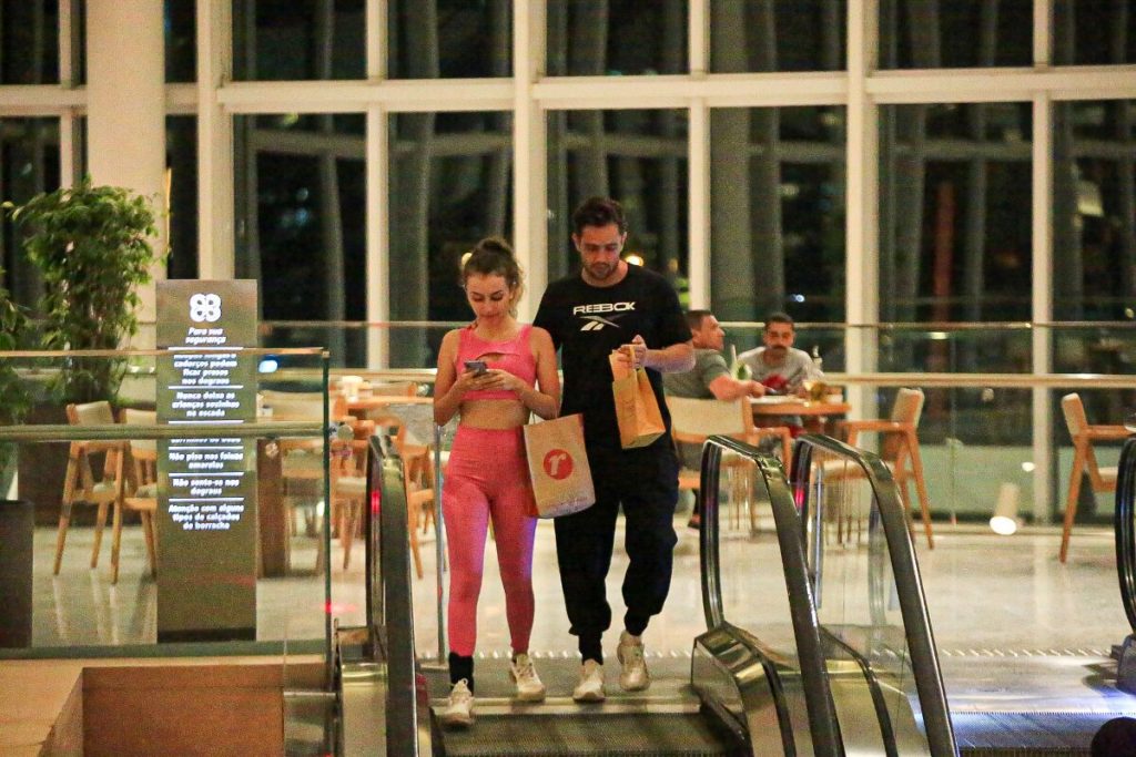 Rafael Cardoso de camisa preta e a namorada, Vivian Linhares, de top e legging rosa na escada rolante