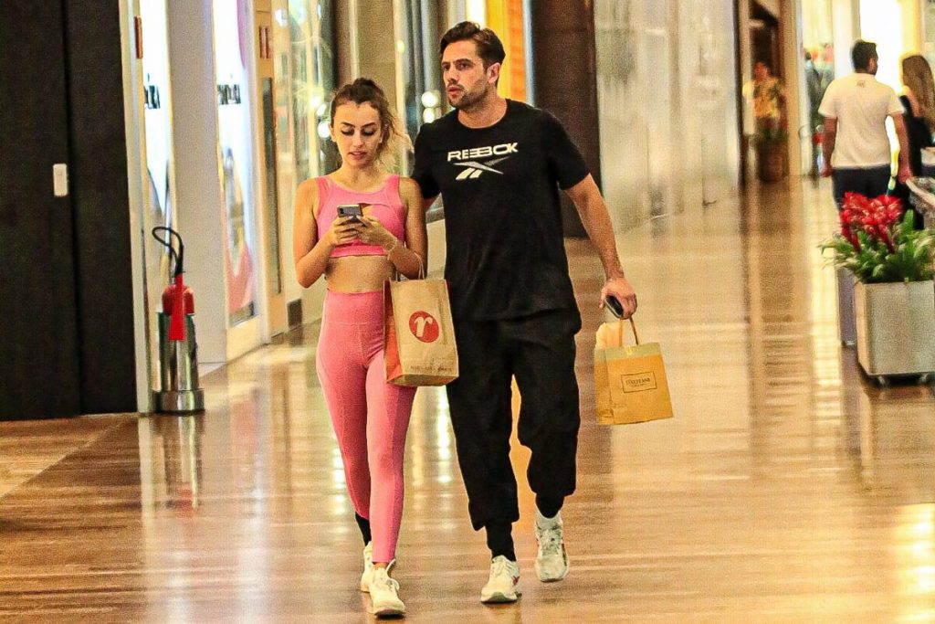 Rafael Cardoso de camisa preta e a namorada, Vivian Linhares, de top e legging rosa na escada rolante caminhando no corredor do shopping