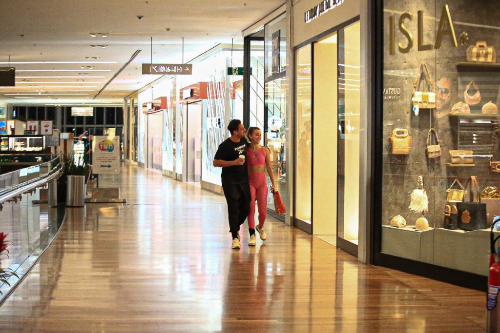 Rafael Cardoso de camisa preta e a namorada, Vivian Linhares, de top e legging rosa na escada rolante caminhando no corredor do shopping