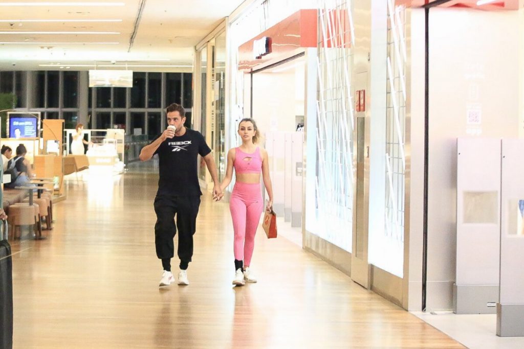 Rafael Cardoso de camisa preta e a namorada, Vivian Linhares, de top e legging rosa na escada rolante caminhando no corredor do shopping