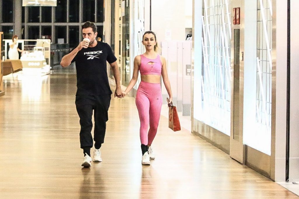 Rafael Cardoso de camisa preta e a namorada, Vivian Linhares, de top e legging rosa na escada rolante caminhando no corredor do shopping