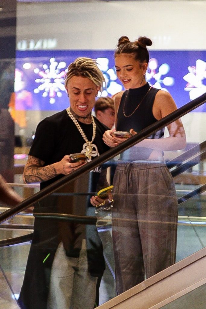 Mel Maia e MC Cabelinho de mãos dadas, escoltados por um segurança, ambos de calça bege e blusa preta 