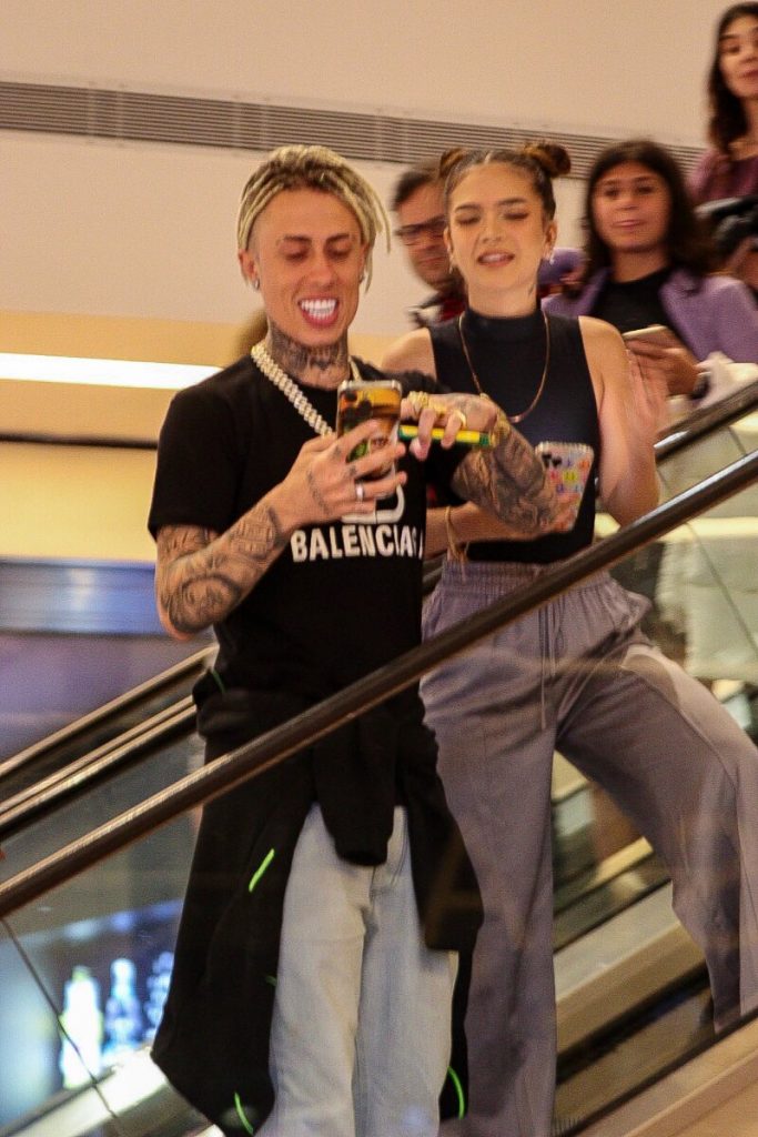 Mel Maia e MC Cabelinho de mãos dadas, escoltados por um segurança, ambos de calça bege e blusa preta 