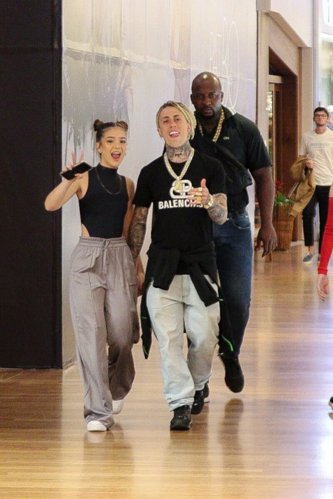 Mel Maia e MC Cabelinho de mãos dadas, escoltados por um segurança, ambos de calça bege e blusa preta 