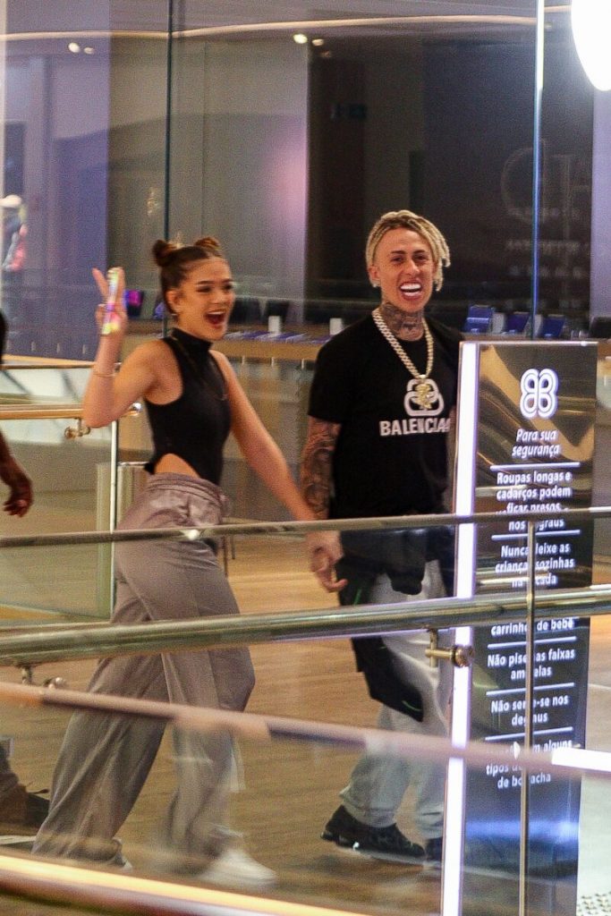 Mel Maia e MC Cabelinho de mãos dadas, escoltados por um segurança, ambos de calça bege e blusa preta 