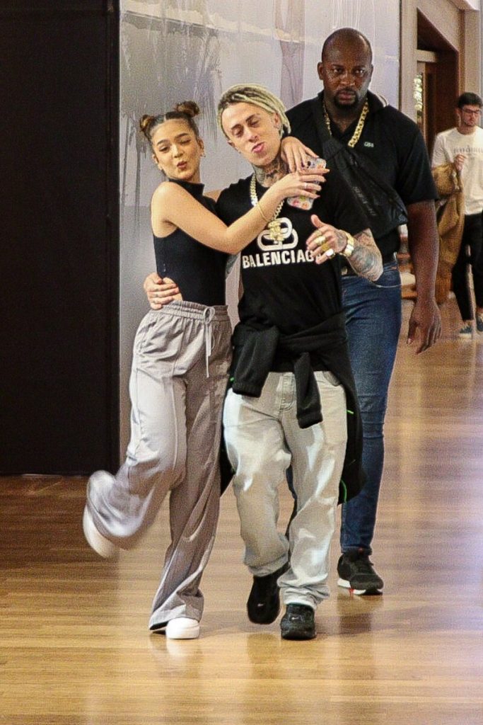 Mel Maia e MC Cabelinho de mãos dadas, escoltados por um segurança, ambos de calça bege e blusa preta 