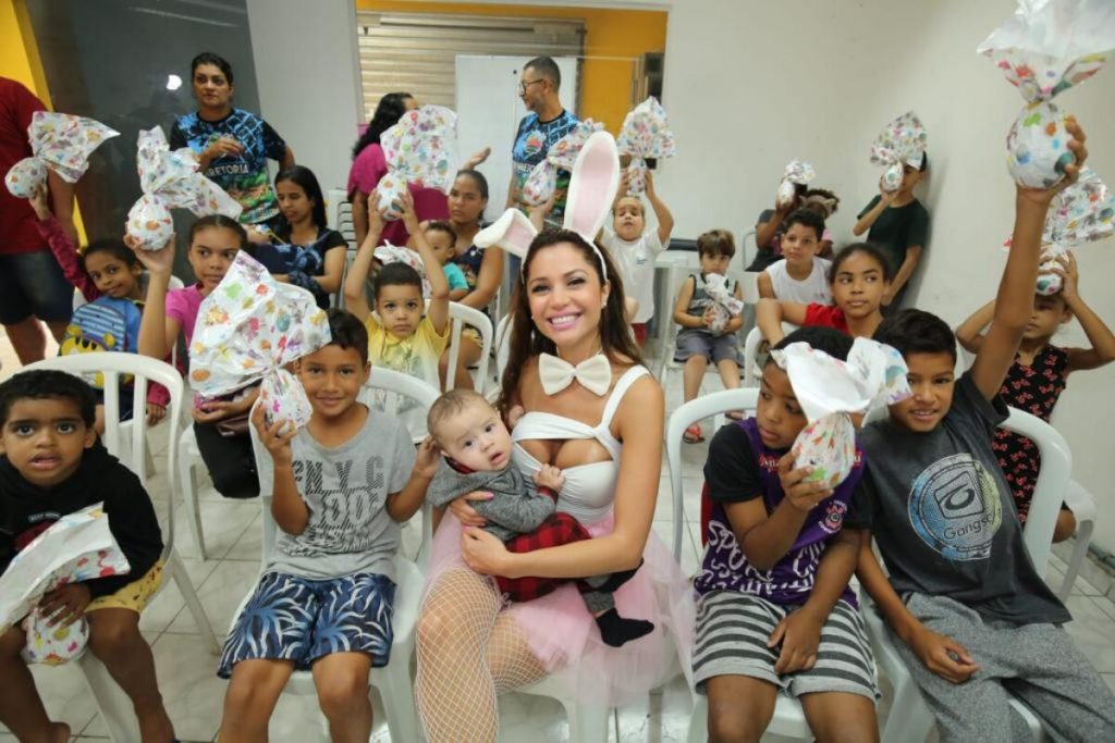 Maria Mellilo vestida de coelhinha, segurando um bebê no colo e rodeada por crianças 