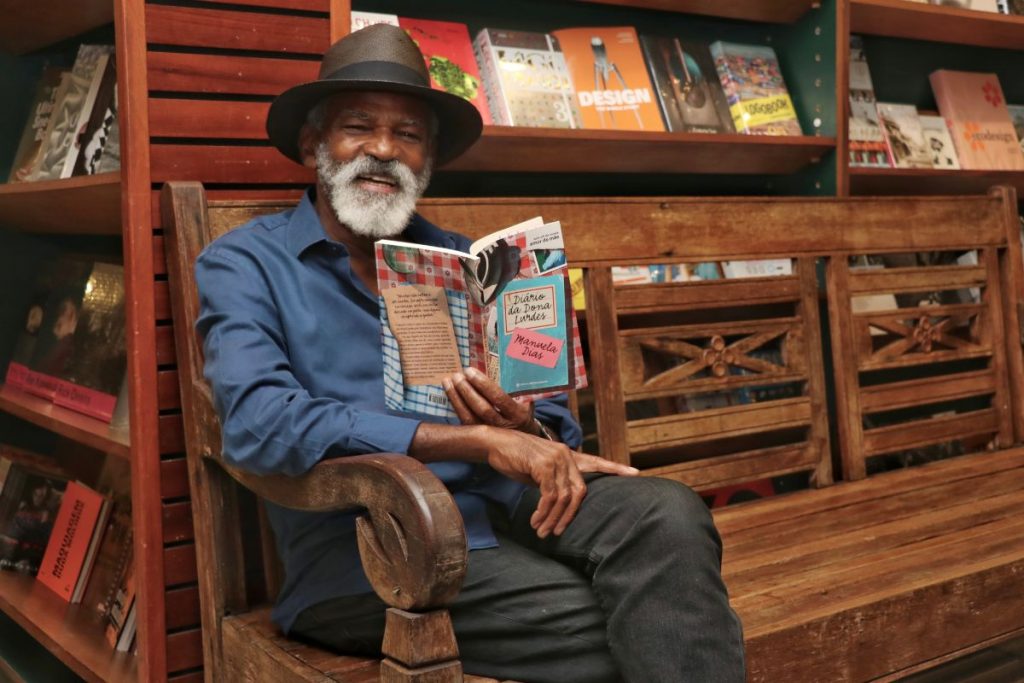 Antônio Pitanga sentado no banco, lendo o lançamento Diário de Dona Lurdes