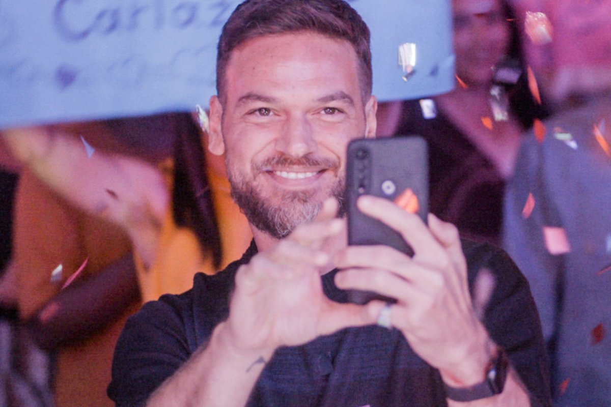 Theo (Emilio Dantas) gravando vídeo com celular e sorrindo em Vai na Fé
