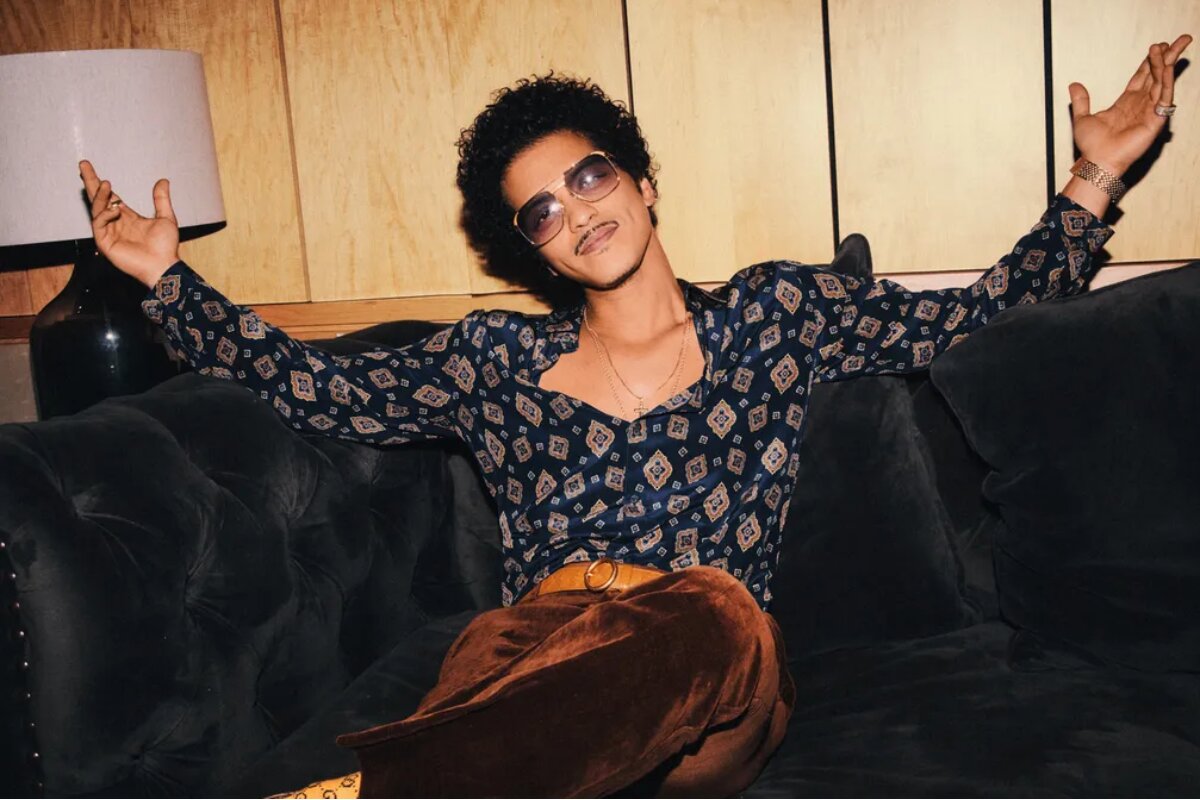 Bruno Mars sentado, de braços abertos e óculos escuros