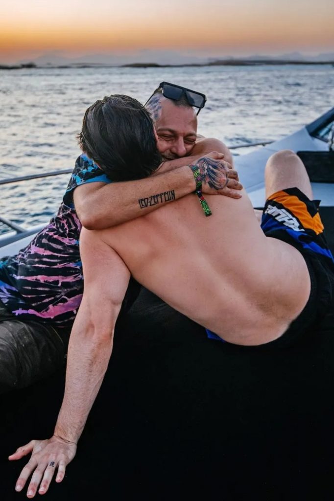 Alok de costas sem camisa abraçado ao pai, em um barco 