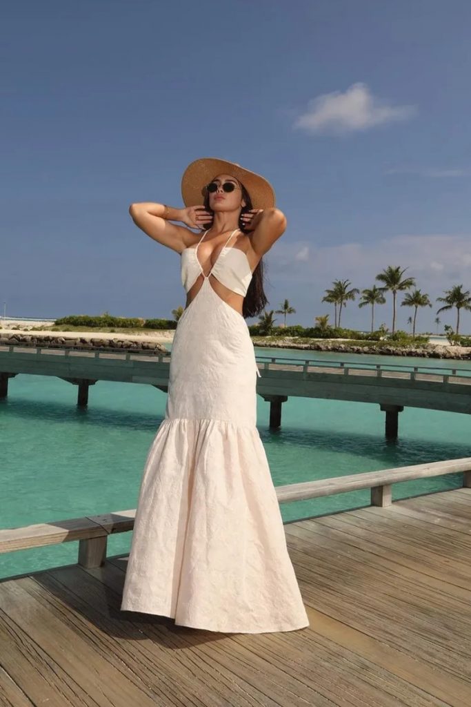 Larissa Santos de vestido branco de recortes e chapéu, sentada em um deck