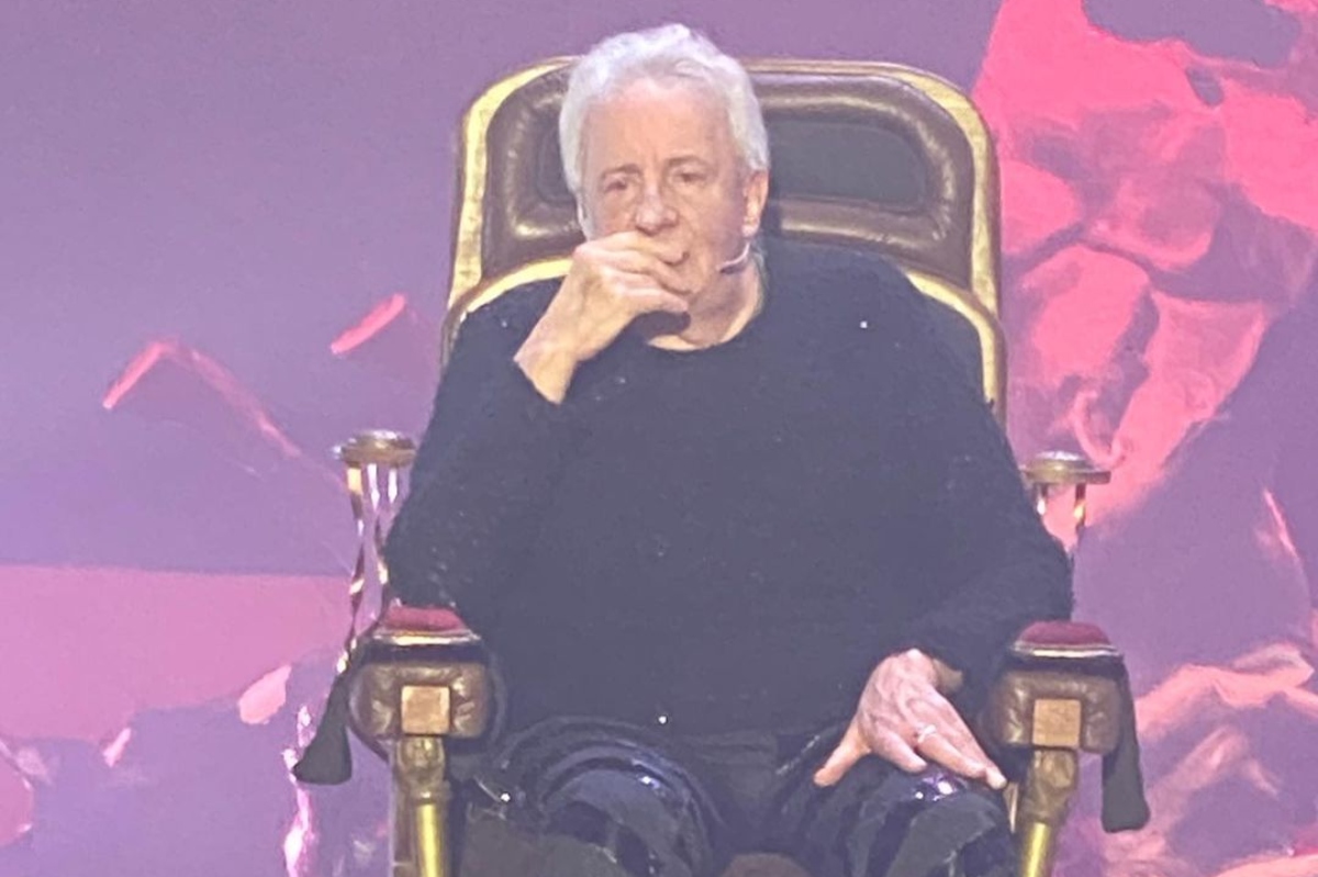 Marco Nanini sentado em uma cadeira grande, de camisa preta, com a mão no queixo