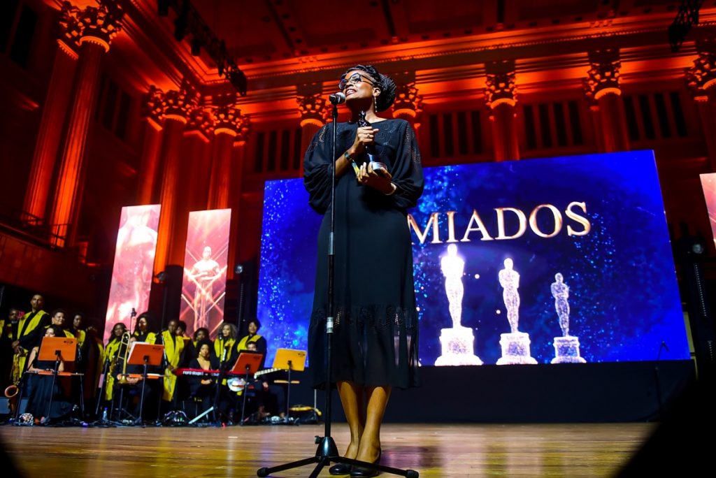 Djamila Ribeiro de vestido preto de manga comprida, segurando troféu, falando ao microfone