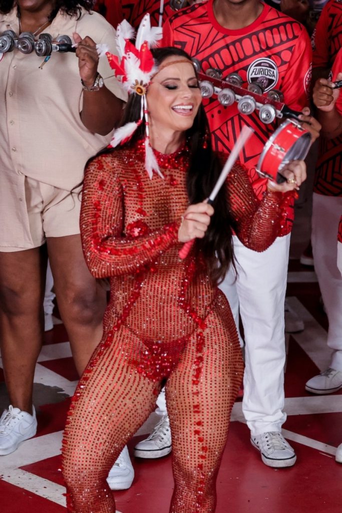 Viviane Araújo de macacão de meia arrastão vazado vermelho, biquíni vermelho tocando tamborim