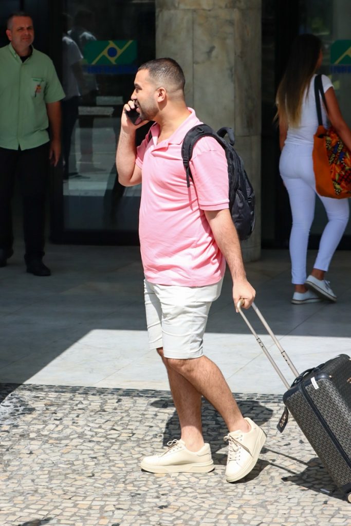 Gil do Vigor de camisa polo rosa e bermuda nude, falando ao telefone, empurrando mala, com mochila mas costas  