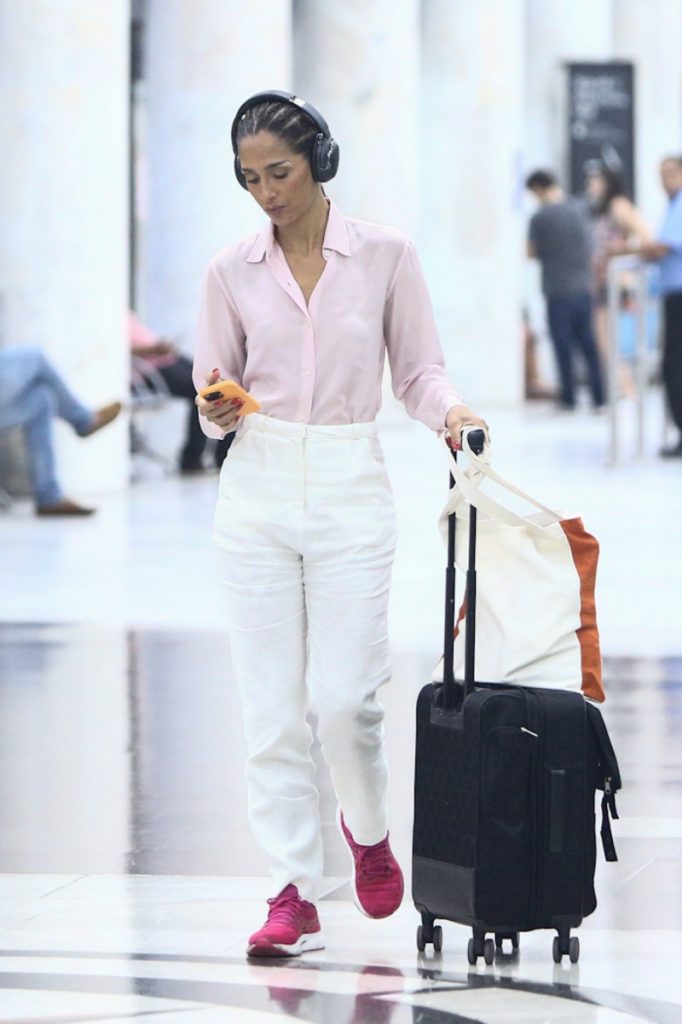 Camila Pitanga de calça branca, camisa social de manga cor de rosa fone de ouvido, com mala, desembarcando no aeroporto 