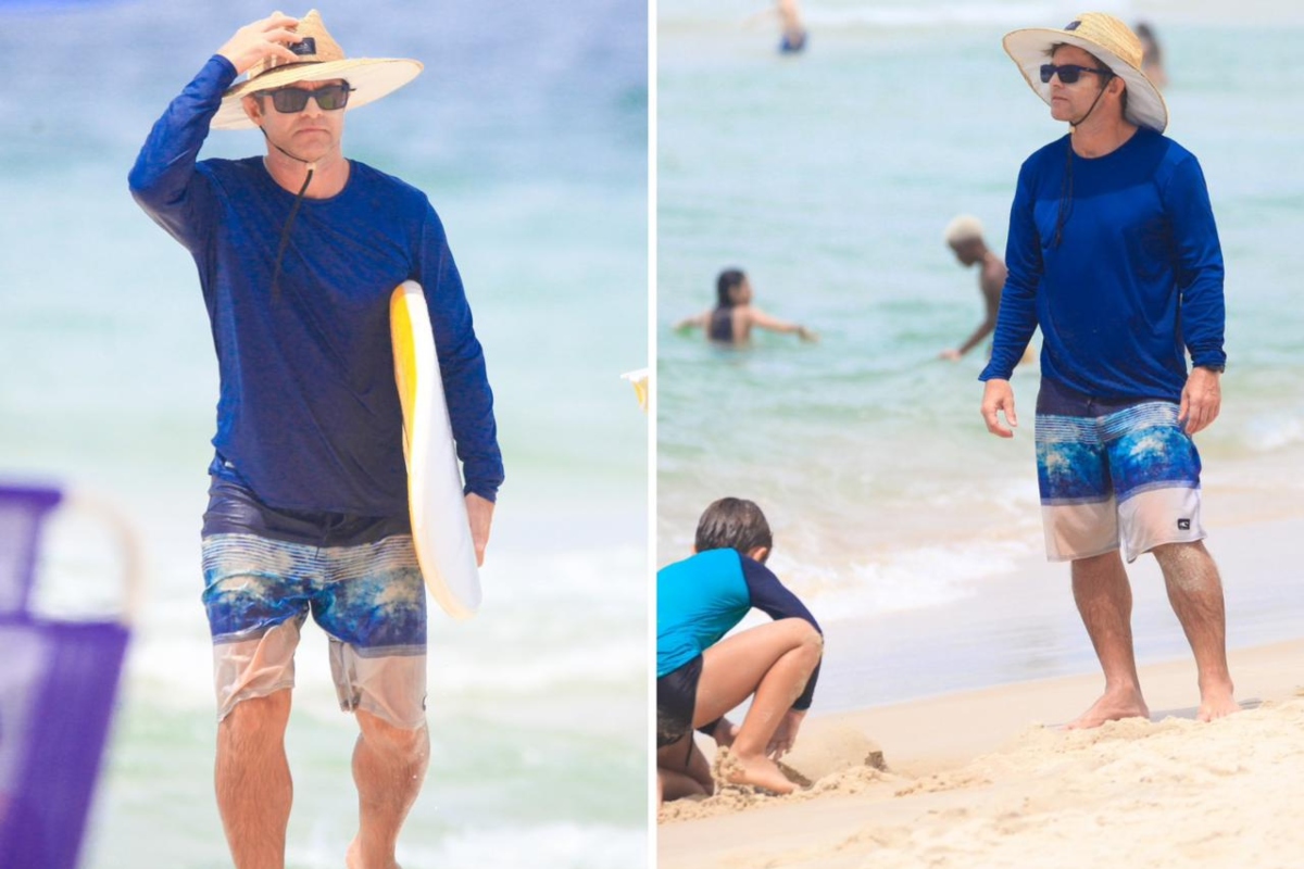 Claudio Heinrich de chapéu de palha, óculos escuros, camisa de manga azul e bermuda estampada, na praia