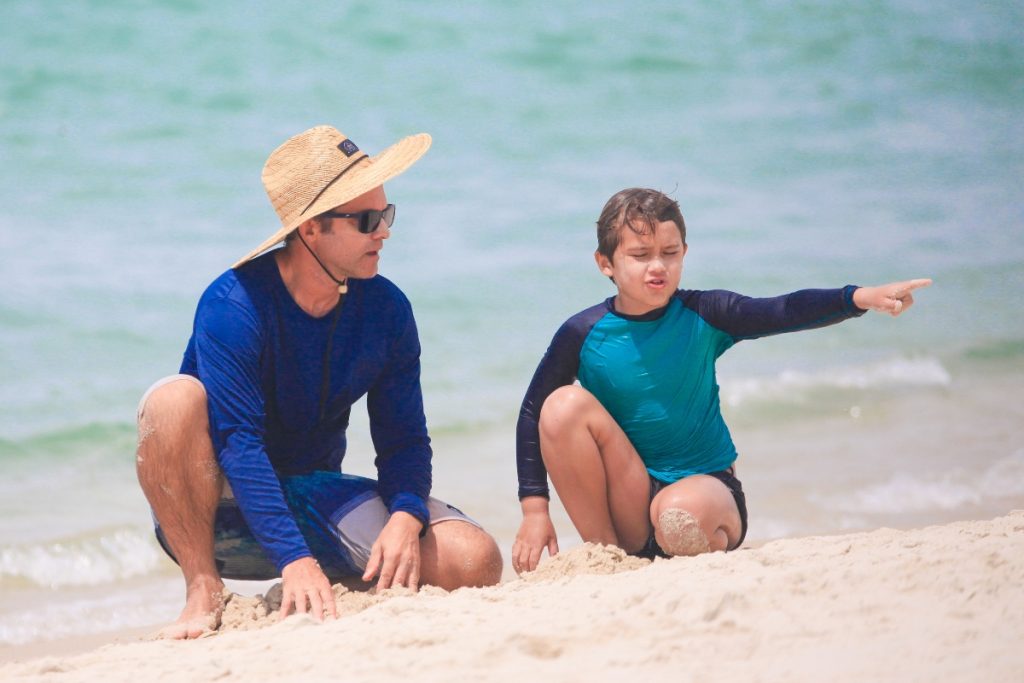 Claudio Heinrich de chapéu de palha, óculos escuros, camisa de manga azul e bermuda estampada, na praia com o filho