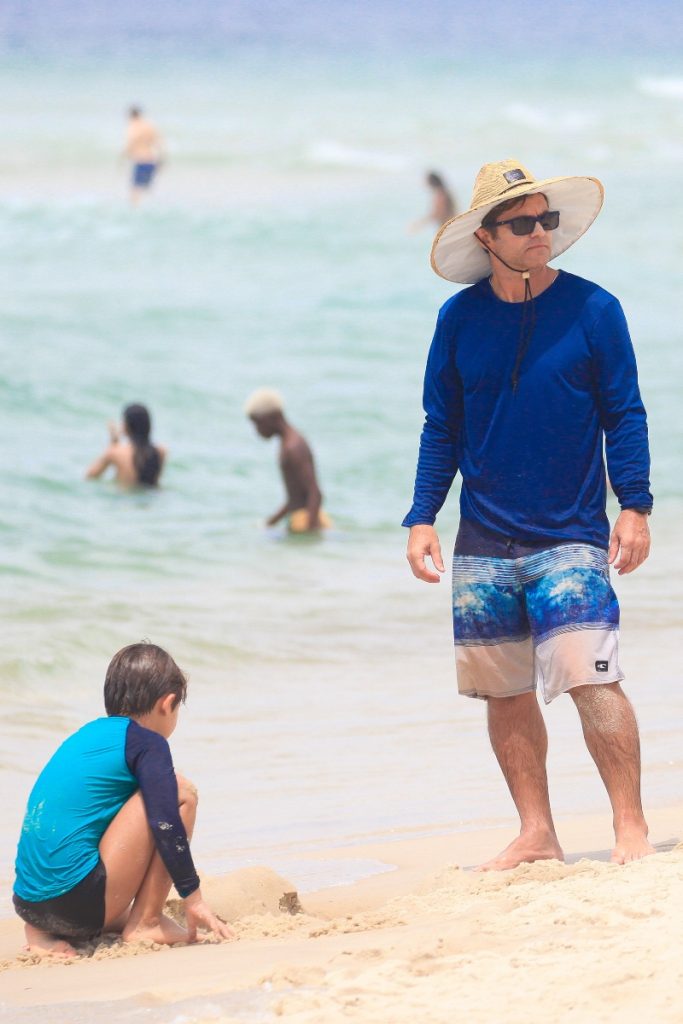 Claudio Heinrich de chapéu de palha, óculos escuros, camisa de manga azul e bermuda estampada, na praia com o filho