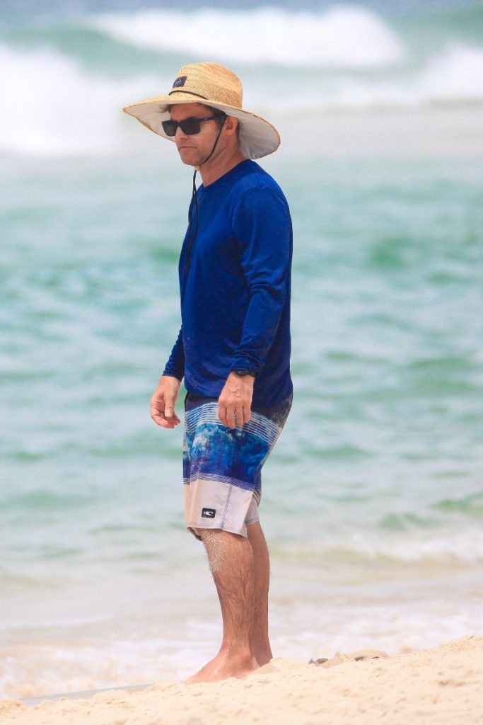 Claudio Heinrich de chapéu de palha, óculos escuros, camisa de manga azul e bermuda estampada, na praia 