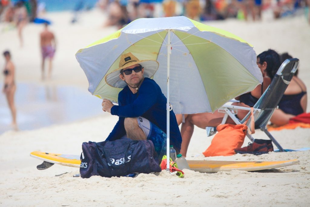 Claudio Heinrich de chapéu de palha, óculos escuros, camisa de manga azul e bermuda estampada, na praia 