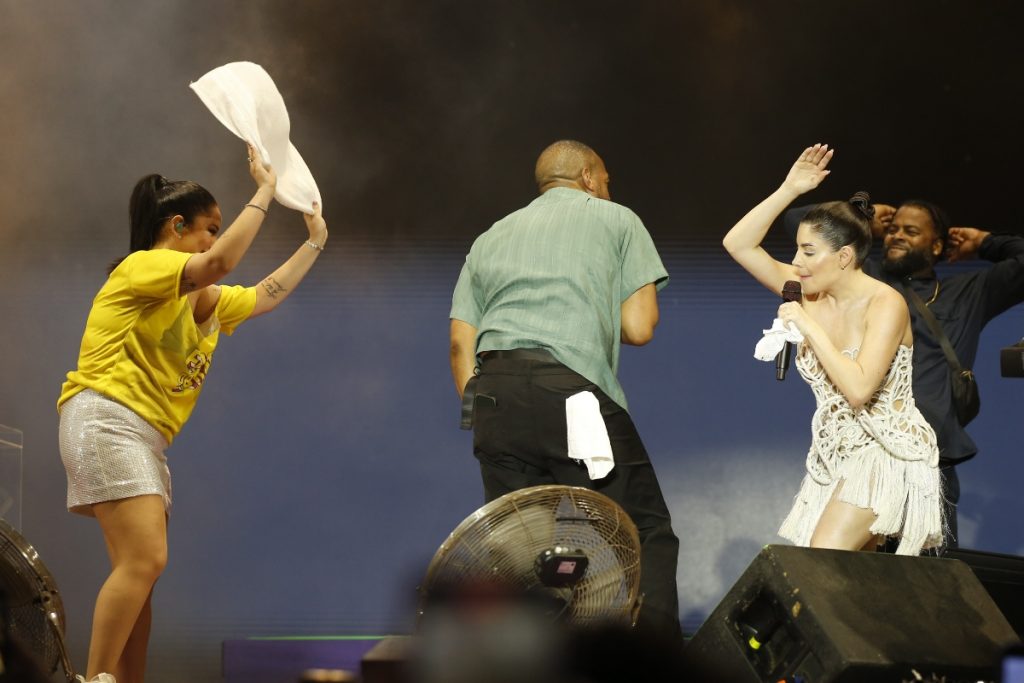 Marlon Wayans de camisa verde e calça jeans, de costas no palco com Gkay, de vestido curto branco e Mari Fernandez de short branco e camisa amarela
