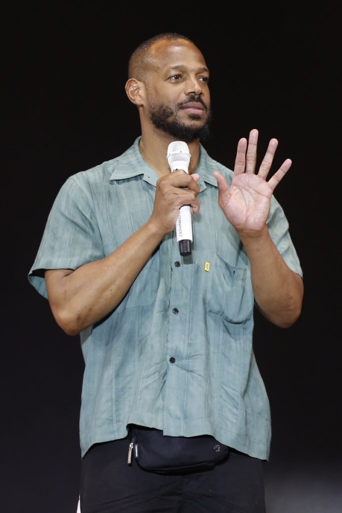 Marlon Wayans de camisa verde e calça jeans, no palco, falando ao microfone 