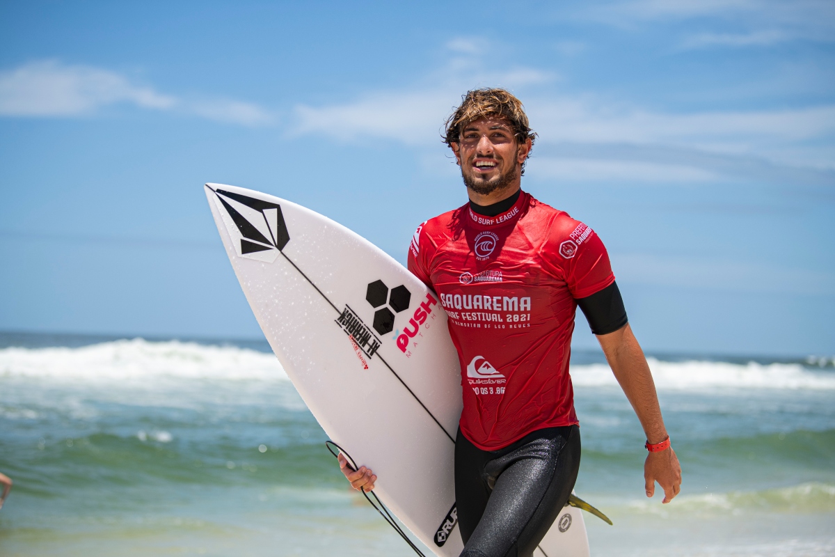 João Chianca de camisa vermelha, segurando prancha de surf, na praia