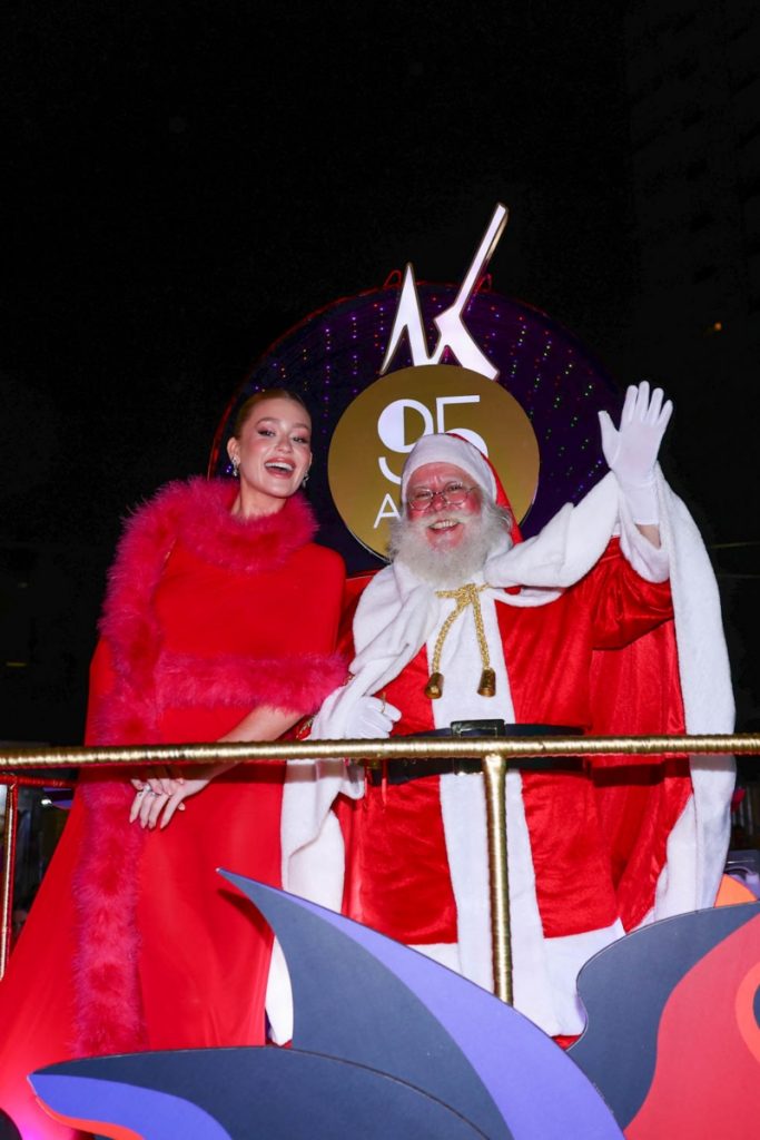 Marina Ruy Barbosa de vestido  vermelho transparente e buá de pelúcia sentada ao lado do Papai Noel 