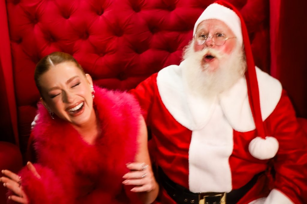 Marina Ruy Barbosa de vestido  vermelho transparente e buá de pelúcia sentada ao lado do Papai Noel 