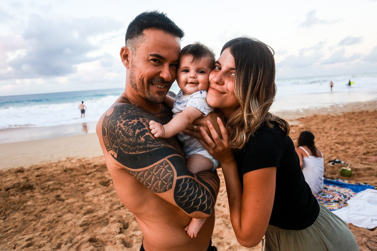 Paulo Vilhena sem camisa, bermuda preta, segurando a a filha, Manoela, abraçado a mulher, Maria Luiza, na praia